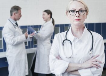 Kollarını bağlamış ön planda duran gözlüklü kadın doktor ve arka planda meslektaşları.