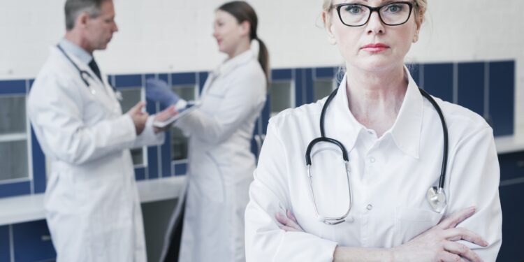 Kollarını bağlamış ön planda duran gözlüklü kadın doktor ve arka planda meslektaşları.