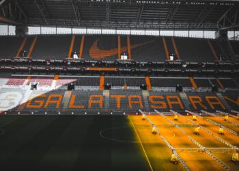 Sarı-kırmızı renkli tribünleri ve çim sahası görünen boş bir futbol stadyumu.
