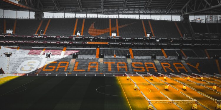 Sarı-kırmızı renkli tribünleri ve çim sahası görünen boş bir futbol stadyumu.