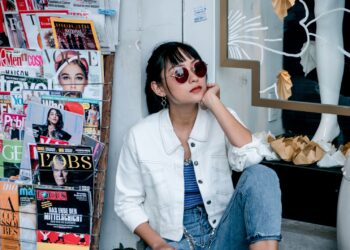 Güneş gözlüklü genç bir kadın sokakta magazin dergilerinin önünde oturuyor.