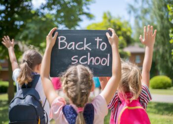 Sırt çantalı üç küçük kız arkası dönük şekilde ellerinde Back to School yazılı kara tahta tutuyor.