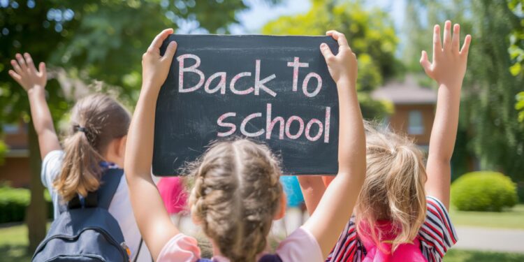Sırt çantalı üç küçük kız arkası dönük şekilde ellerinde Back to School yazılı kara tahta tutuyor.