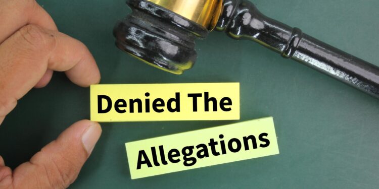 Bir elin tuttuğu sarı blok üzerinde 'Denied The Allegations' yazısı ve mahkeme tokmağı bulunuyor.