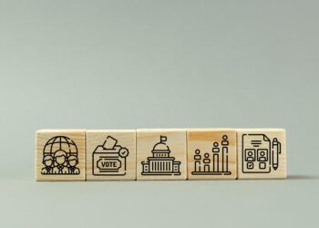 Gri zemin üzerinde oy sandığı, küre ve kamu binası simgeleri bulunan ahşap bloklar.